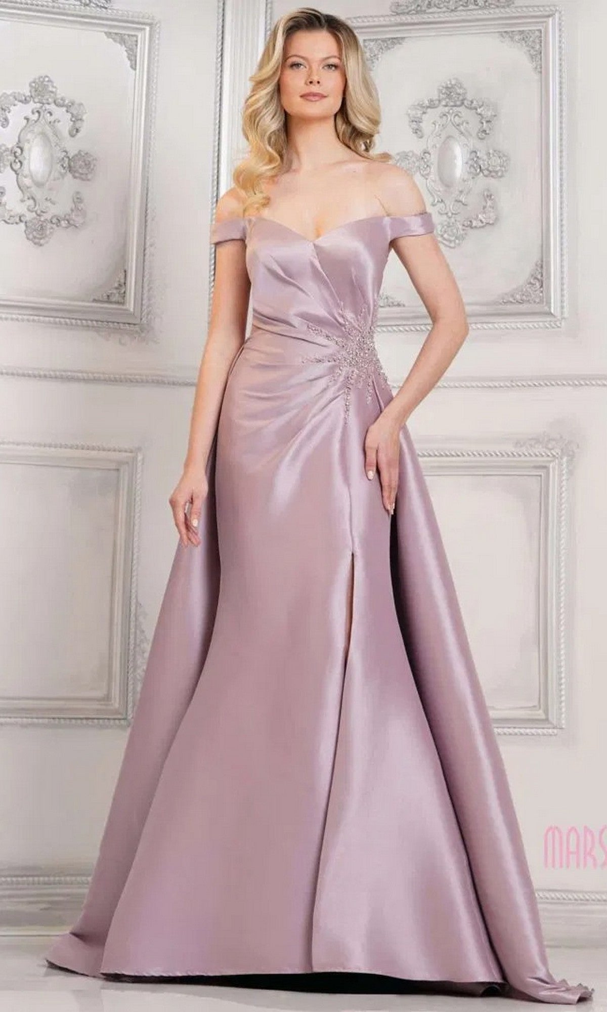 A woman stands in an ornate, light-colored room wearing the Marsoni MV1310 long formal dress in dusty rose, featuring an off-shoulder neckline, mermaid silhouette, front slit, and waist embellishment.